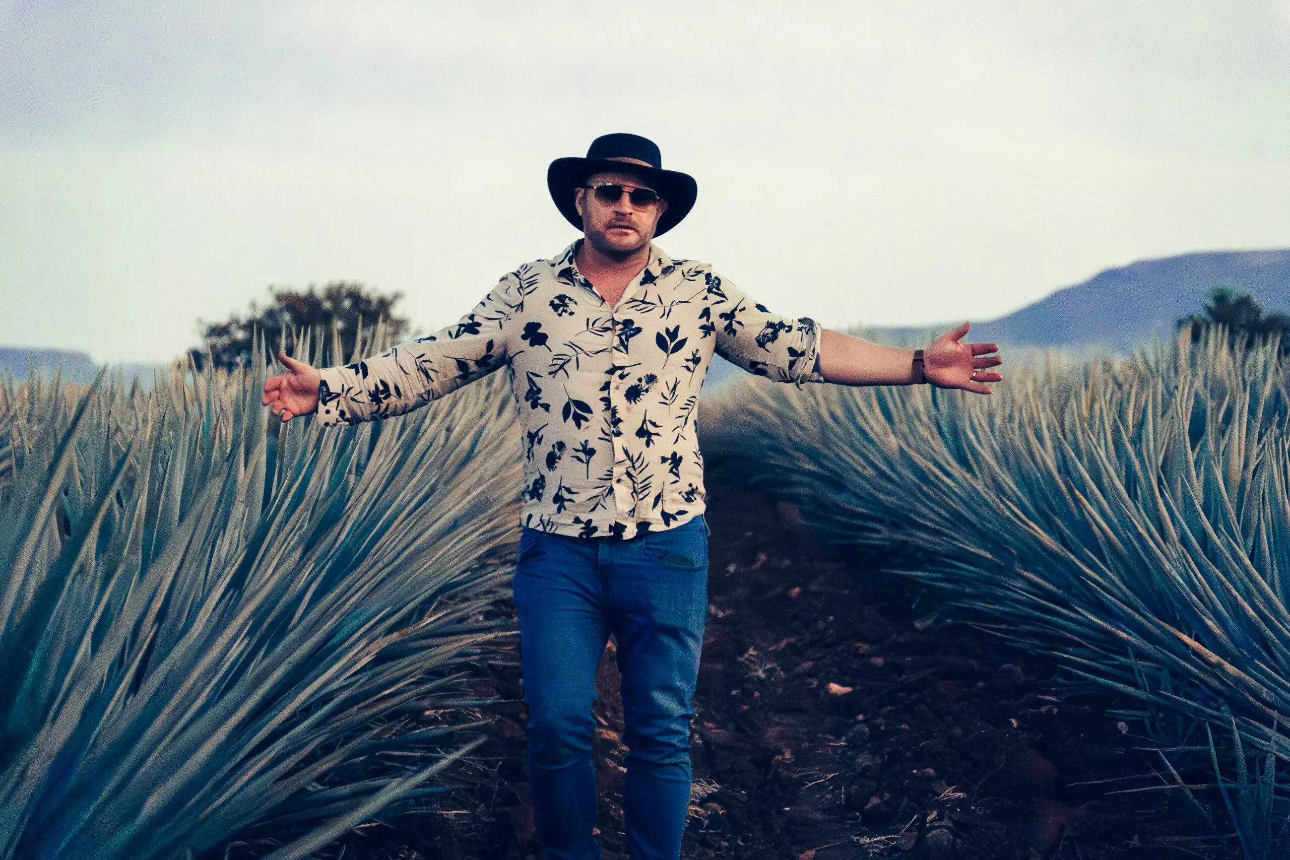 Man posing with arms open in an agave field, wearing a hat and patterned shirt.