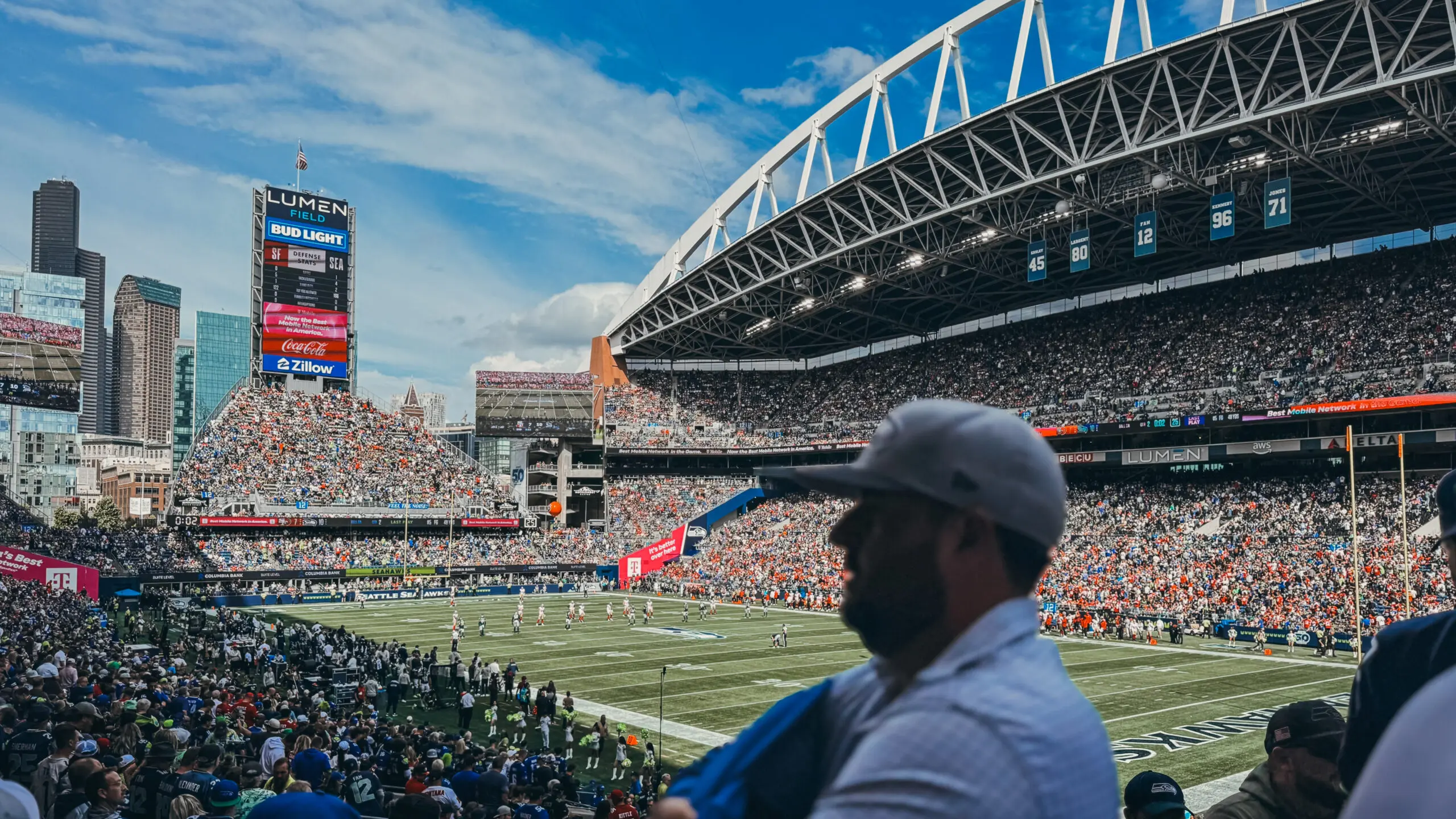 A man wearing sunglasses watches a football game in a packed stadium.