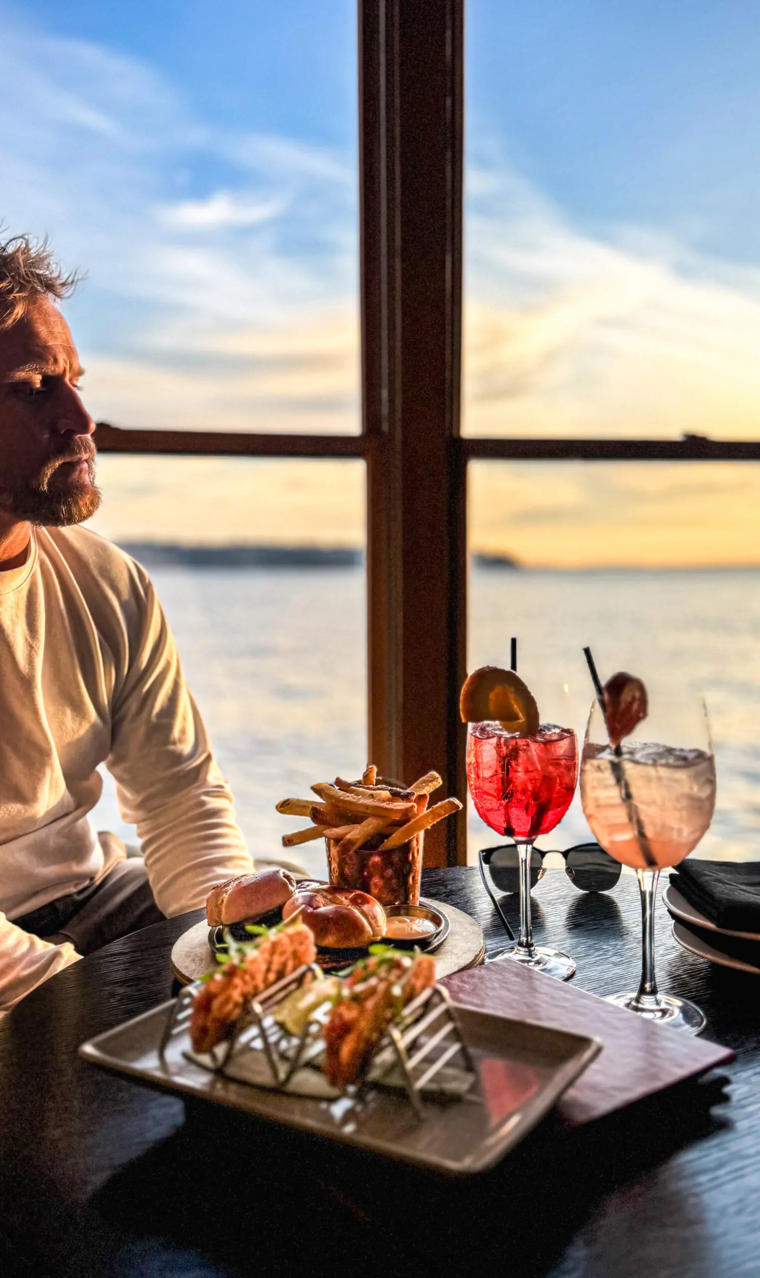 Man enjoying a meal and drinks by a sunset window.