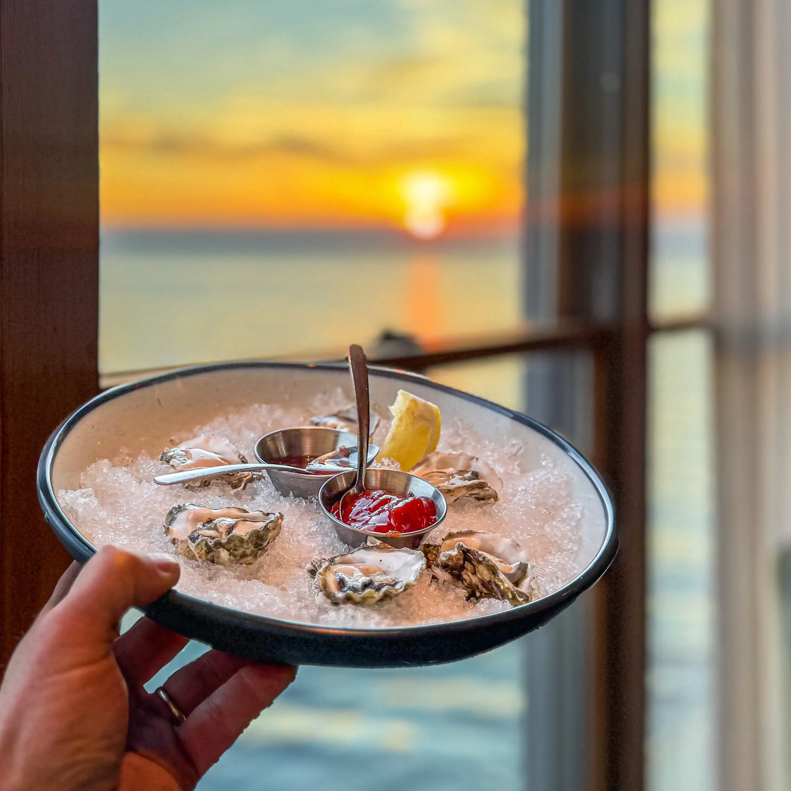 Hand holding a bowl of dessert with nuts and cherry at sunset by the window.
