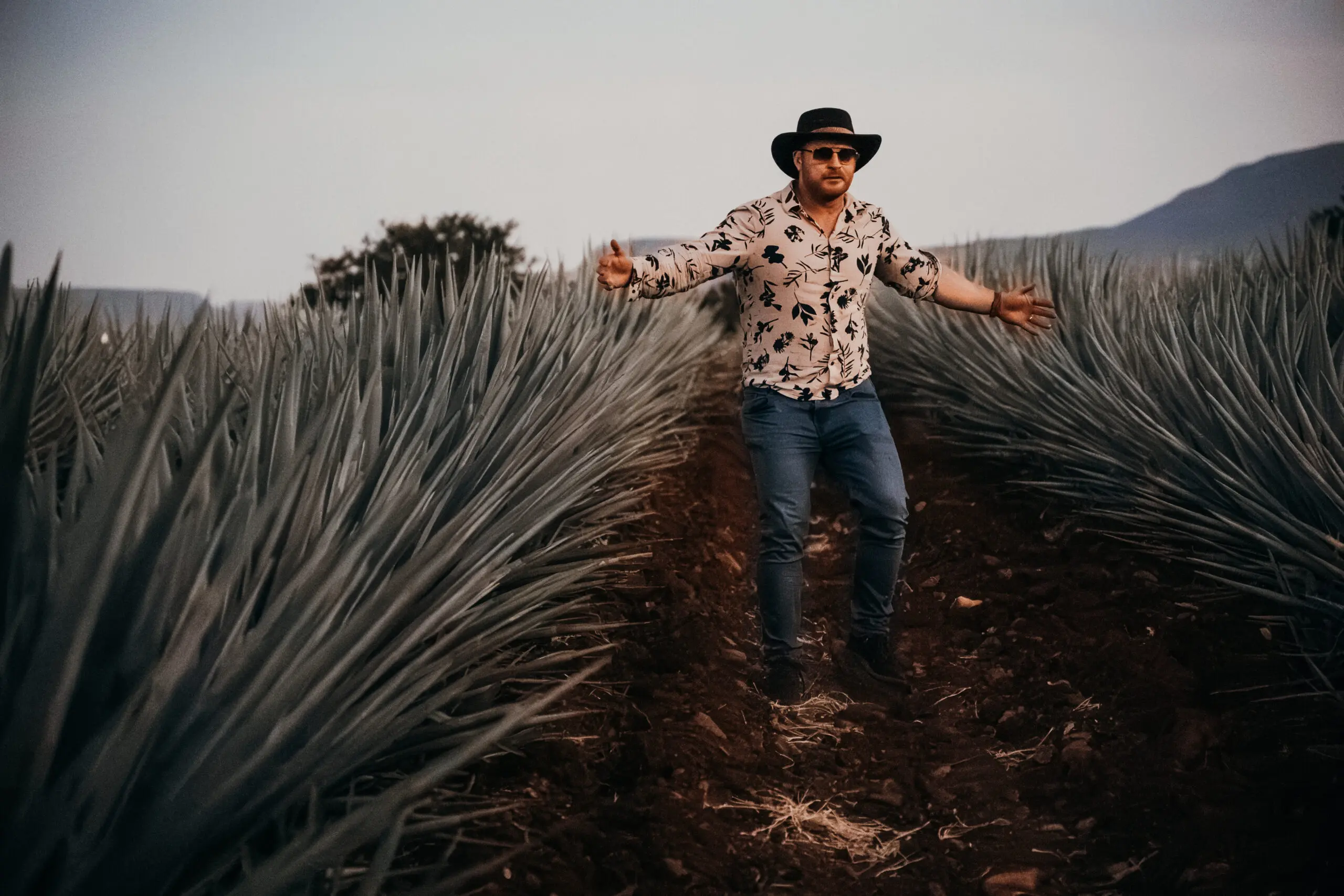 Man in a patterned shirt and hat walking through tall plants outdoors.