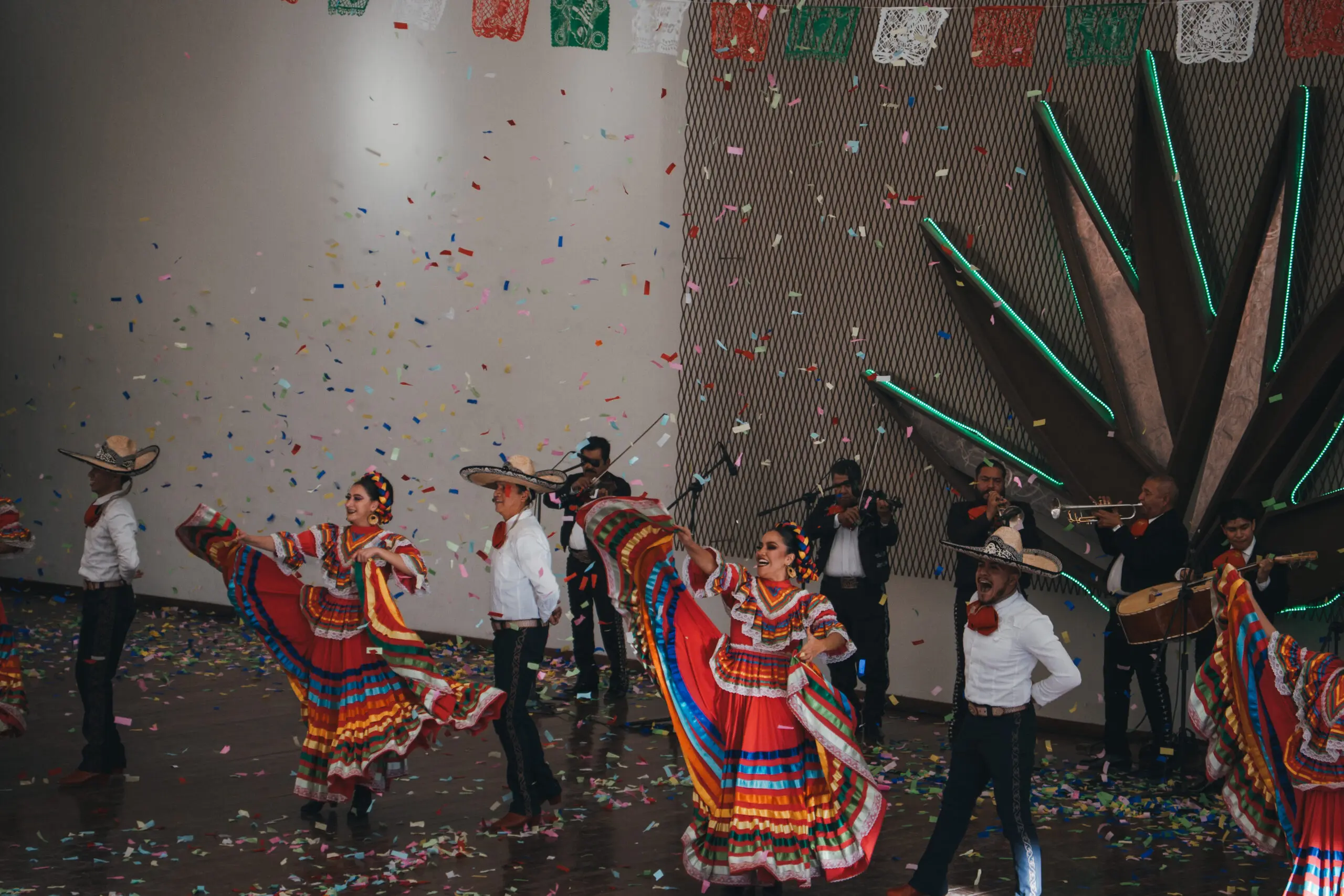 Dancers in colorful traditional attire perform beside a climbing wall indoors.