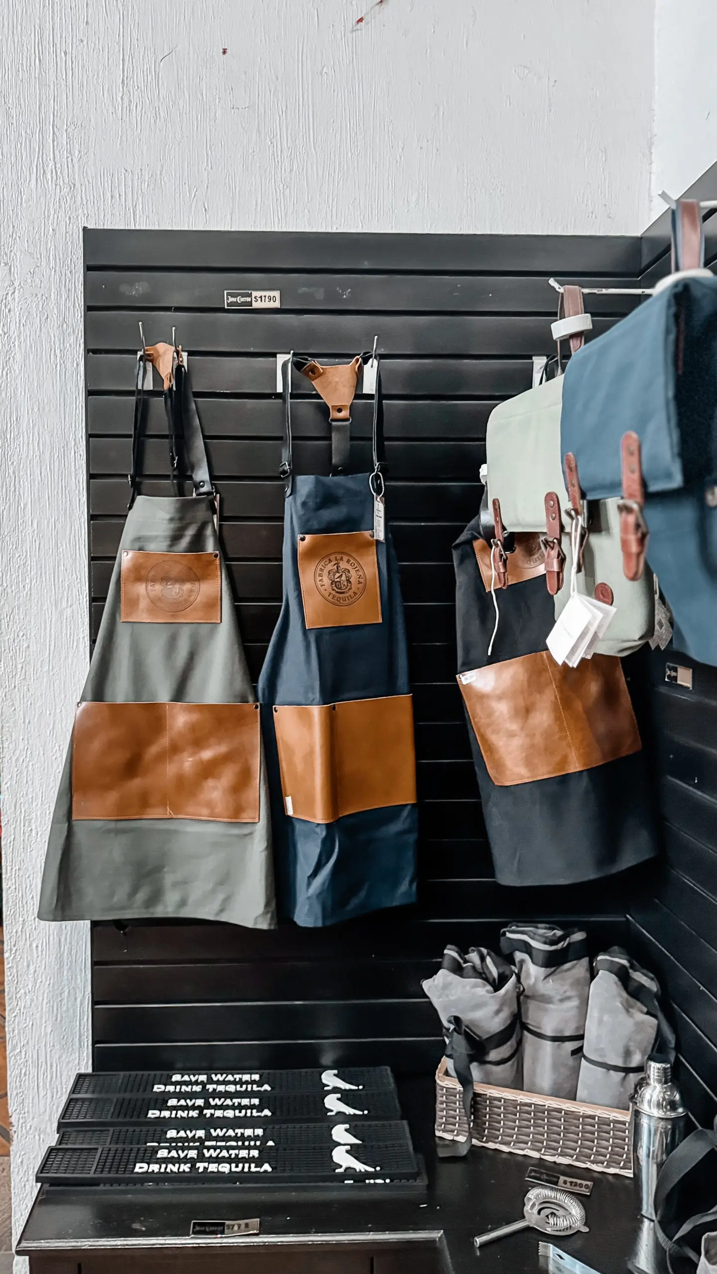 Two aprons hanging on hooks against a black wall, featuring leather pockets and straps.
