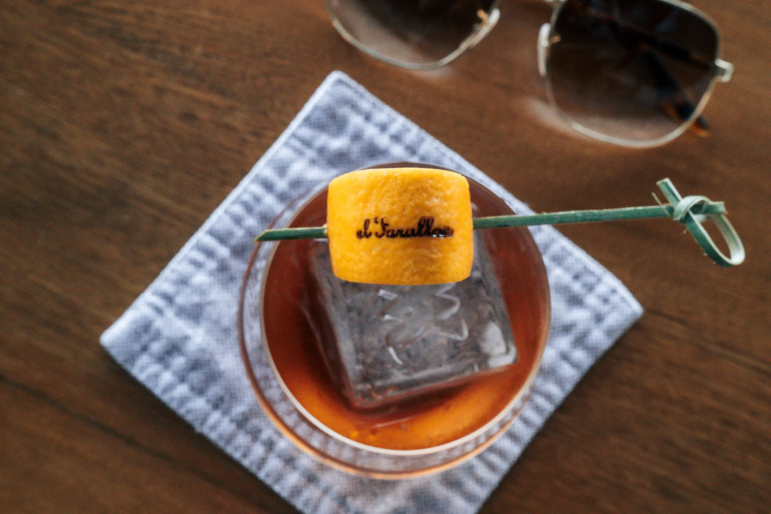 A cocktail with a large ice cube and a toasted marshmallow garnish on a wooden table.