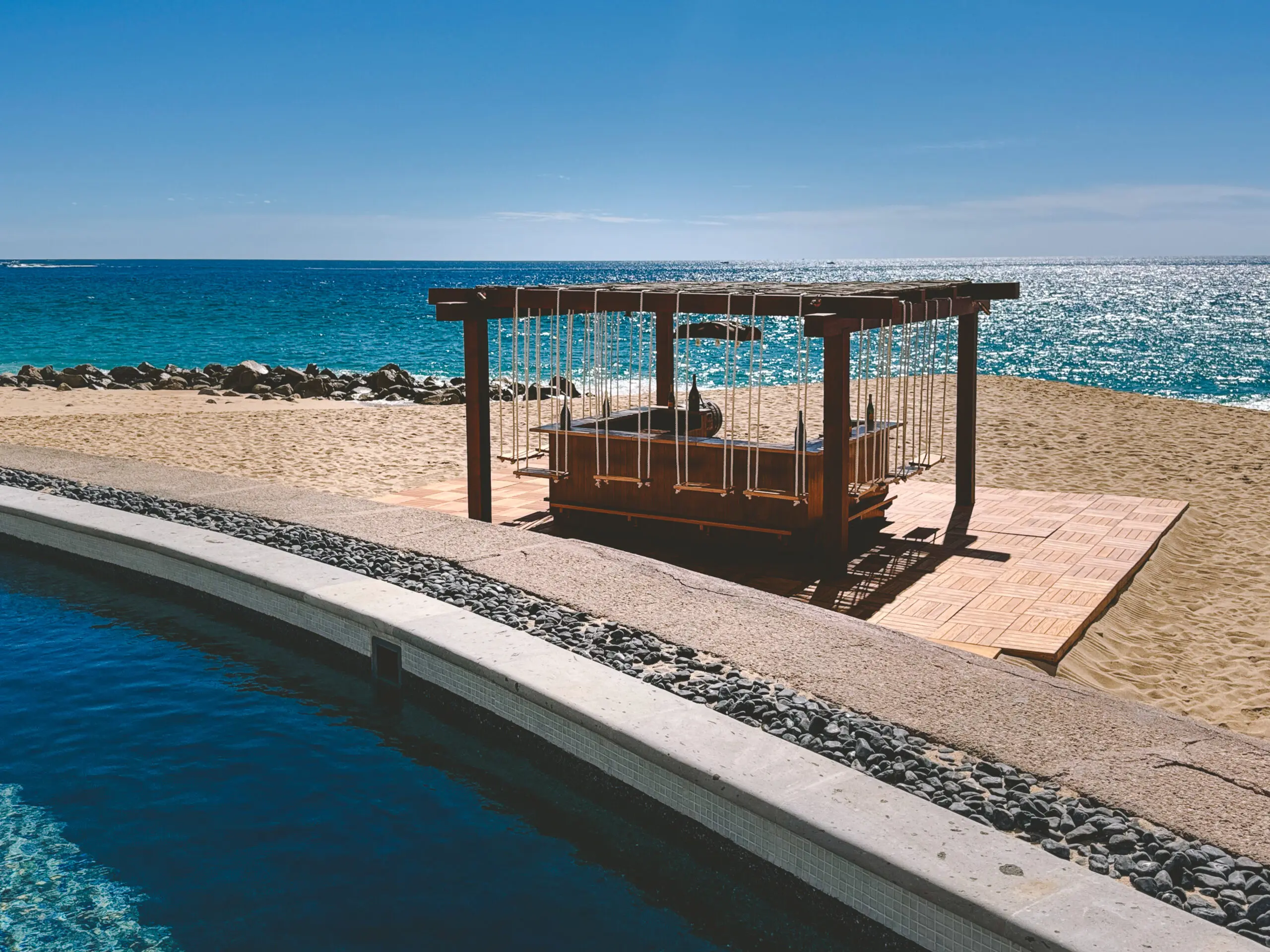 A shaded wooden pergola on a sandy beach near a curved pool under a clear blue sky.