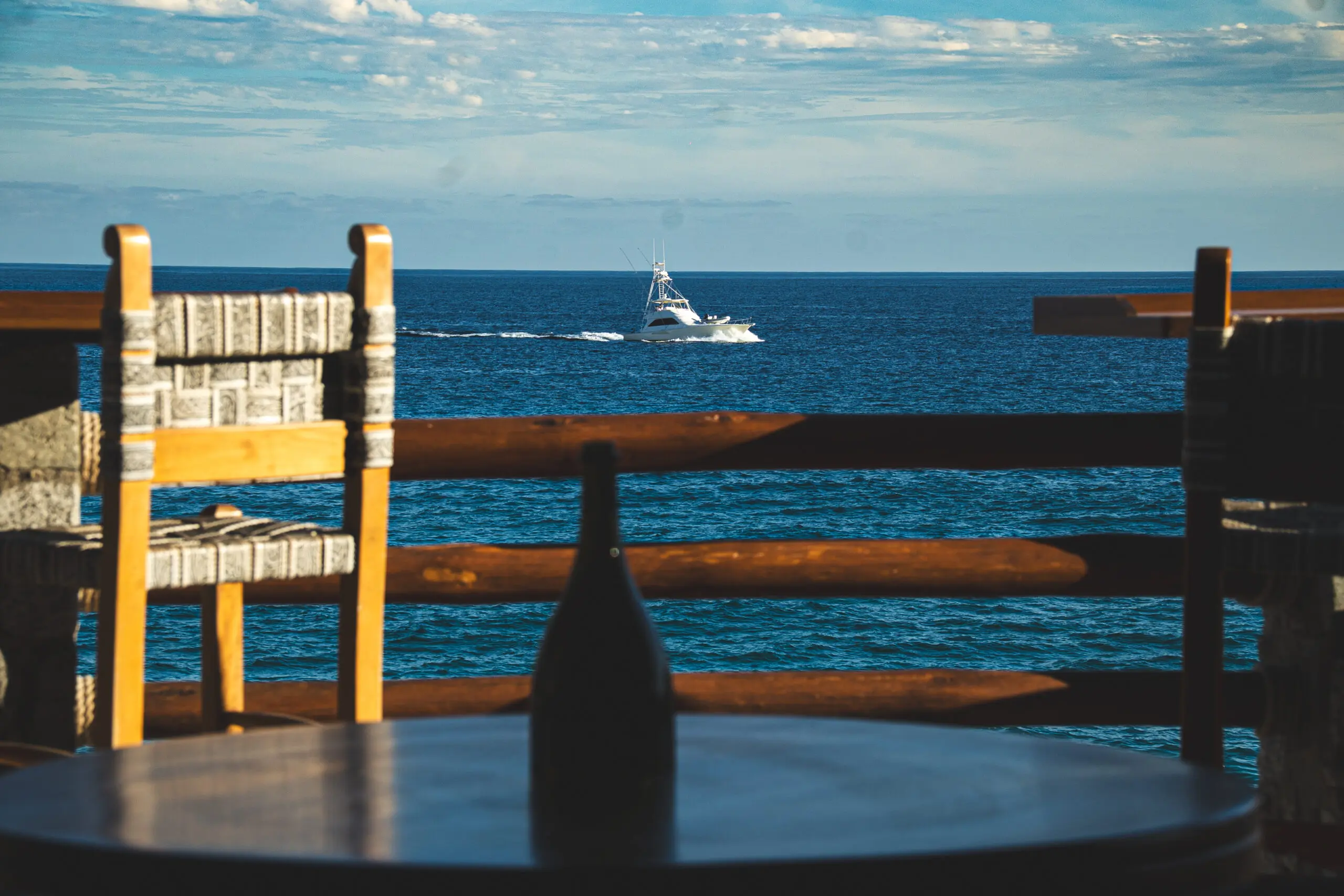 View of a boat sailing on the ocean from a shaded porch with wooden furniture.