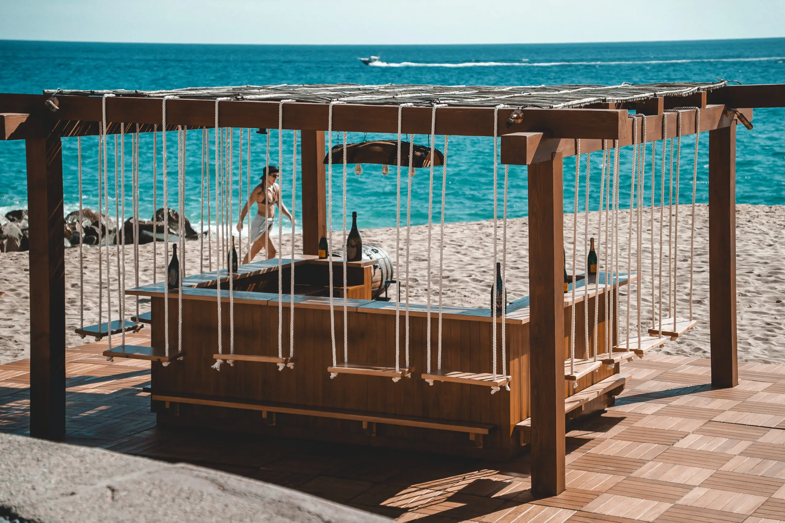 People relaxing on swing seats by the beach under a wooden canopy.