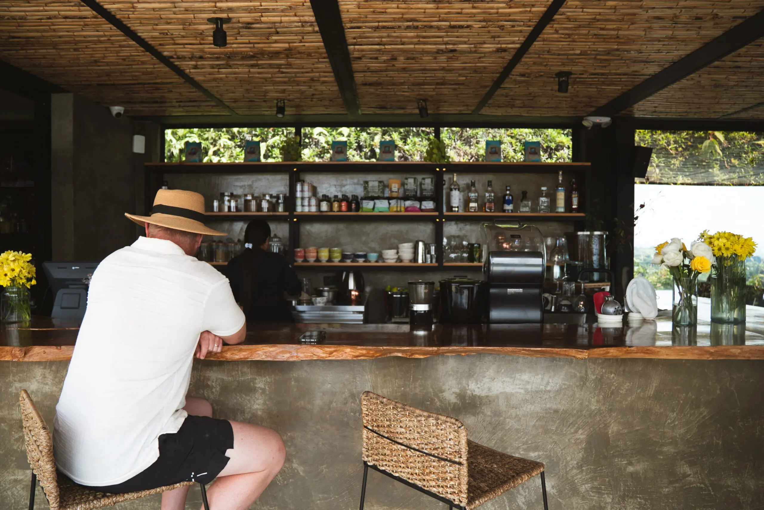 Man sitting alone at a rustic outdoor bar with wooden ceiling and shelves of bottles.