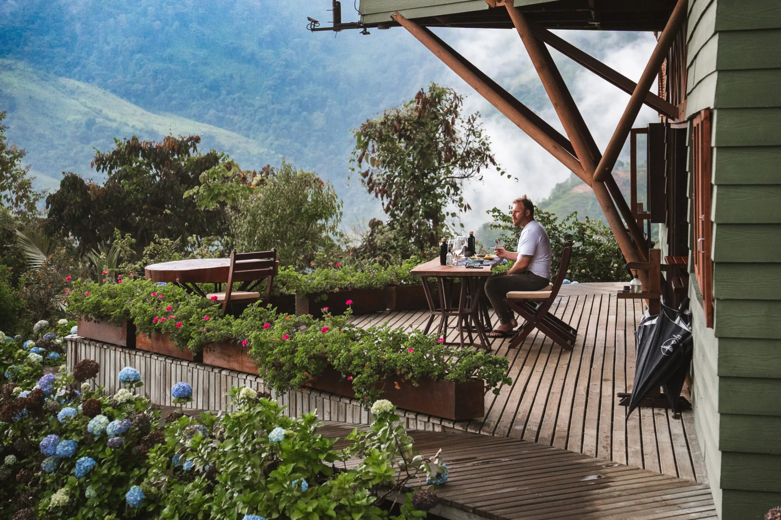 A person sitting alone on a wooden deck surrounded by plants and mountains.