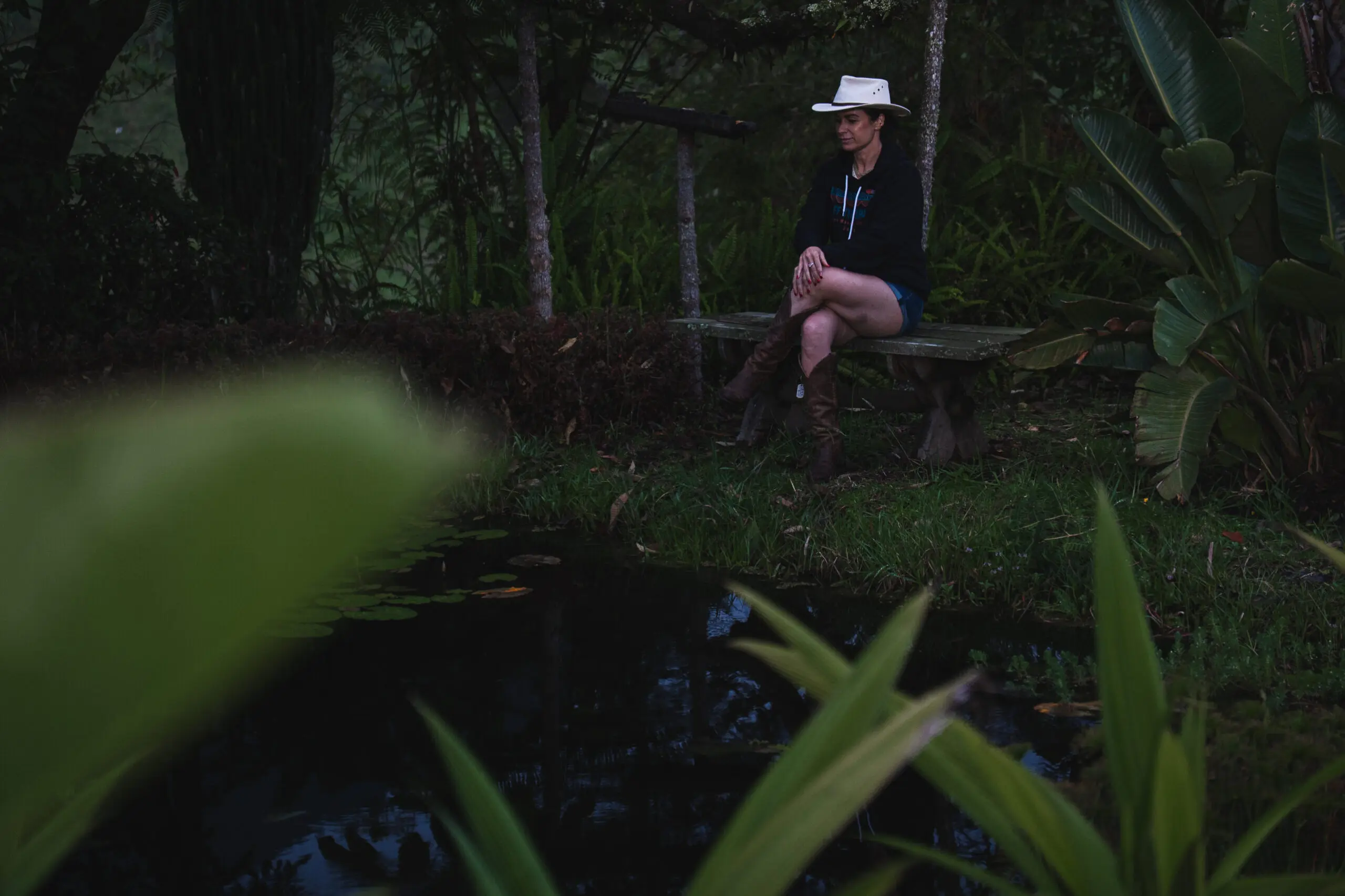 Person in white hat sitting on a log by a dark forest pond at night.