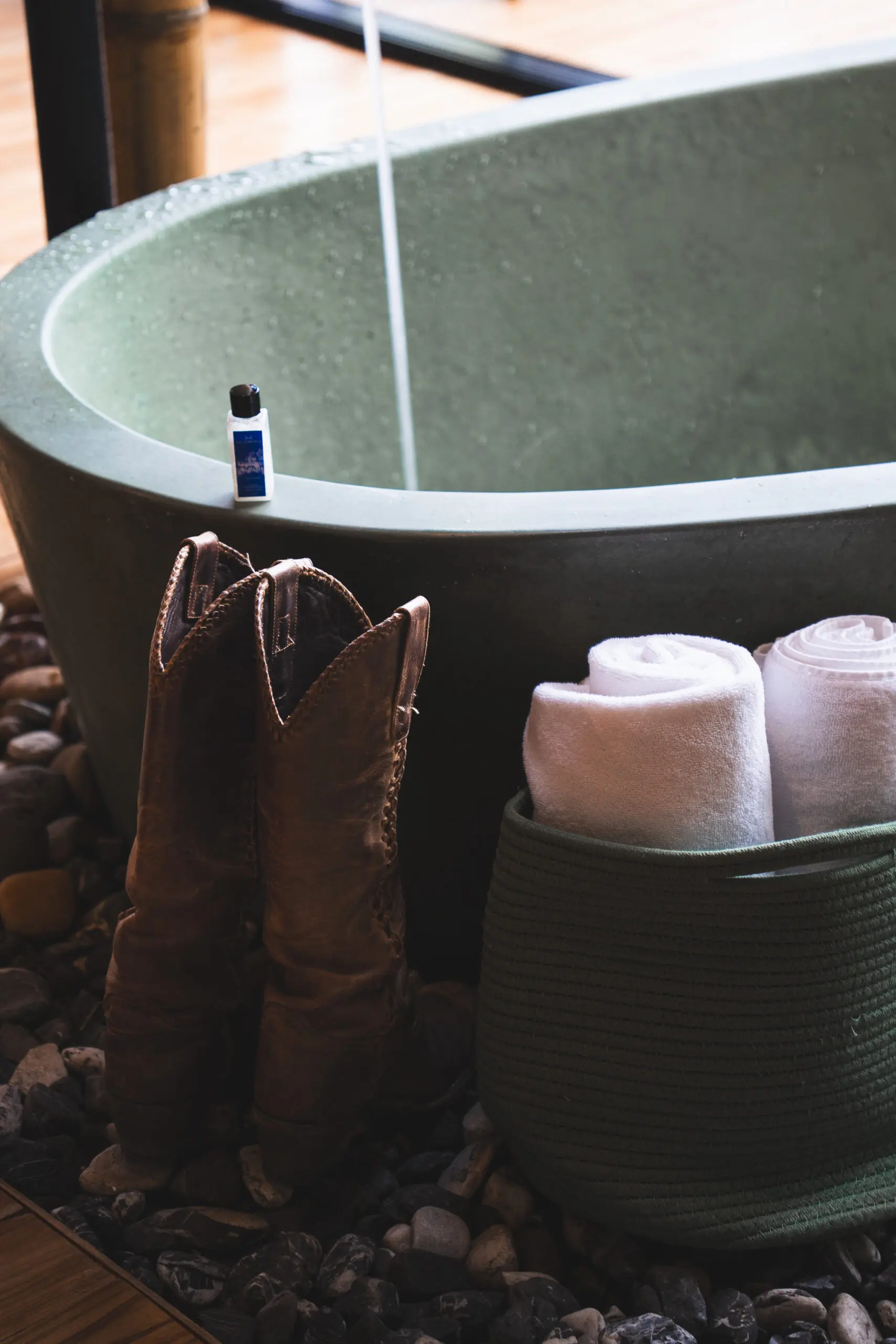 Rustic boots by a stone bathtub with towels and bath oil in a cozy bathroom.