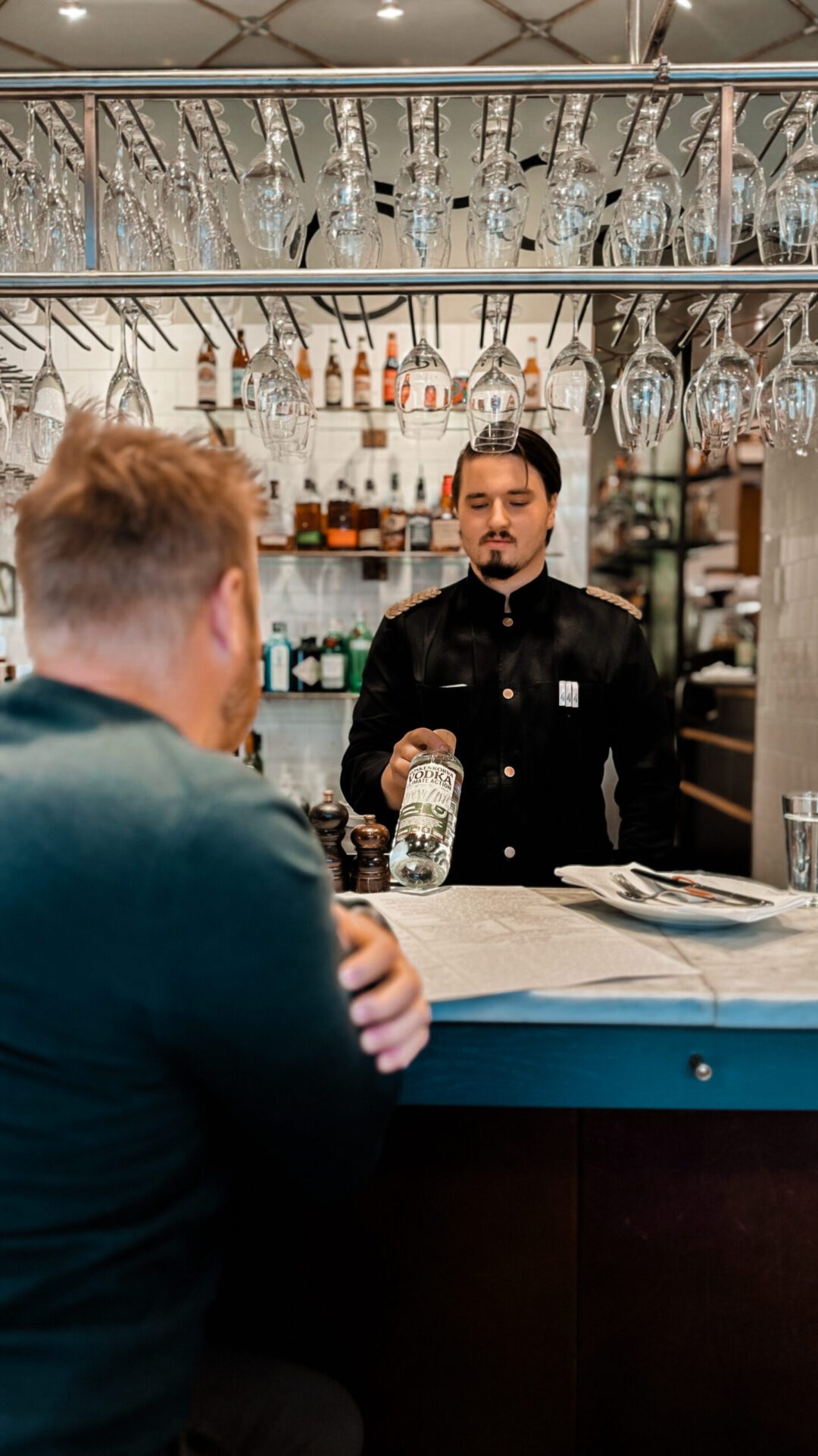 Man dressed in waiter uniform handing a bottle of liquor to a man sitting at the counter